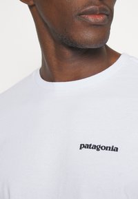 Fehér pamut póló, kerek nyakkivágással és fekete "patagonia" logóval a bal felső mellkasi részen. Puha, sima textúra.
