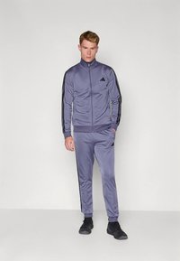 Violets sporta tērps no gluda materiāla, kas ietver rāvējslēdzēja jaku ar melnām svītrām, atbilstošus bikses un melnus sporta apavus.