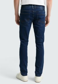Vaqueros ajustados de color azul oscuro con una textura suave, que presentan bolsillos traseros con costuras decorativas y un sutil logo en relieve en la cinturilla.