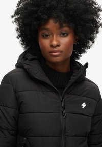 Veste matelassée noire avec fermeture éclair à l'avant et capuche, présentant une texture matelassée et un logo éclair blanc sur le côté gauche de la poitrine.