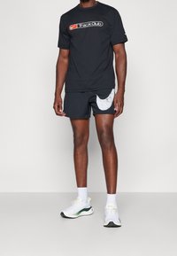 Musta Nike Track Club t-paita logolla, yhdistettynä mustiin shortseihin, joissa on harmaa Nike Swoosh. Valkoiset urheilukengät vihreillä yksityiskohdilla.