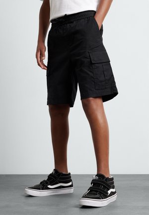Person, der schwarze Cargo-Shorts und schwarze High-Top-Sneaker mit weißen Sohlen trägt, steht mit Händen in den Taschen auf einem grauen Boden.