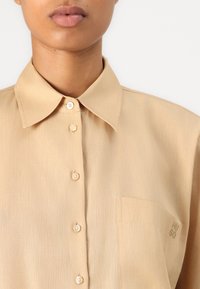 Chemise beige à boutons, fabriquée dans un tissu lisse. Elle comporte un col, une petite poche avant et une broderie de logo discrète sur la poitrine.