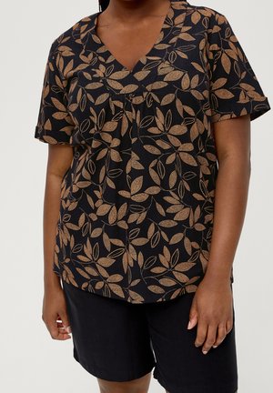 Vrouw die een zwarte V-hals top met bruine bladpatronen draagt en effen zwarte shorts, staand tegen een effen achtergrond.