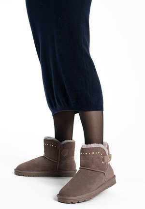 Botas de tobillo de ante marrón con un forro suave y tipo piel de pelo, con tachuelas doradas y un detalle de correa lateral. Las lleva puestas con un vestido de punto azul oscuro.