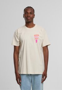 Beige bomullströja med korta ärmar och rundad hals. Har ett livfullt rosa grafiskt tryck på vänster bröstområde. Avslappnad passform.