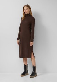 Braunes langärmeliges Kleid mit hohem Kragen und seitlichen Schlitzen, kombiniert mit schwarzen Stiefeletten mit strukturierten Akzenten und dicken Sohlen.