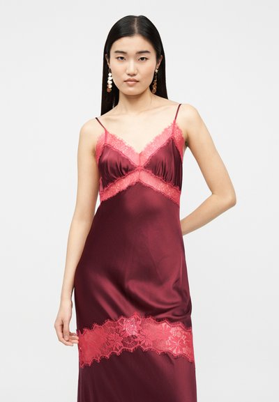 Γυναίκα με μακριά μαύρα μαλλιά φοράει βελούδινο μπορντό φόρεμα χωρίς μανίκια με ροζ δαντέλα στην άκρη και χρυσά σκουλαρίκια με κρεμαστά δίσκους.