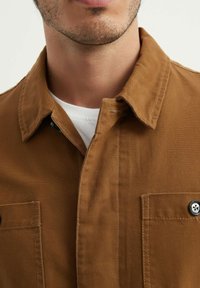 Chemise en coton marron avec un col à boutons, deux poches poitrine et un sous-vêtement blanc visible en dessous. Accents de boutons remarquables sur les poches.