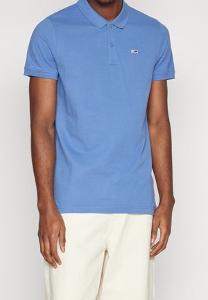 Homme portant un polo bleu avec col et boutons, debout les bras détendus, associé à un pantalon blanc cassé sur un fond uni.