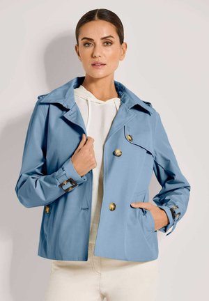 Frau trägt eine hellblaue, zweireihige Jacke mit braunen Knöpfen und Schnallendetails über einem weißen Kapuzenpullover und steht vor einem einfarbigen Hintergrund.