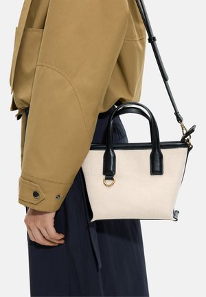 Osoba nosząca beżową kurtkę i ciemne spodnie, niosąca małą beżową torbę typu tote z czarnymi uchwytami i paskiem na ramię.