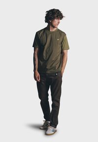 Olijfgroen T-shirt met korte mouwen en een subtiele logo, gecombineerd met donkerbruine broeken en witte sneakers met blauwe en groene accenten.