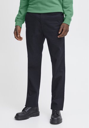 Pantaloni neri a gamba dritta realizzati in tessuto resistente. Presentano un davanti piatto, tasche laterali e una superficie liscia. Indossati con stivali neri.