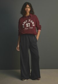 Bordowy sweatshirt z grafiką "HAMPTONS 97 New York" oraz czarnymi spodniami o szerokich nogawkach; luźny krój i miękka tekstura.
