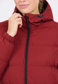 Rød pufferjakke med lynlås og sorte accenter, der har hætte og elastiske manchetter. Stoffet har en glat, tekstureret overflade.