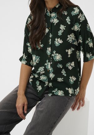 Vrouw draagt een donkergroene blouse met bloemenprint en knopen, met driekwart mouwen en donkergrijze spijkerbroek, zittend op een witte blok.