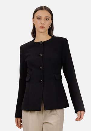 Kvinne iført en tettsittende svart knappet blazer med lange ermer og klafflommer, kombinert med beige bukser, stående mot en hvit bakgrunn.