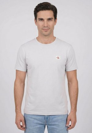 Uomo che indossa una t-shirt a maniche corte grigio chiaro con una piccola toppa con il logo sul petto sinistro e jeans azzurri, in piedi su uno sfondo semplice.