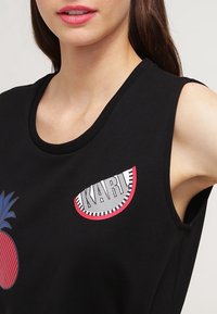 Svart ärmlös topp med en ananasgrafik och en patch med "KARL" i ett randigt mönster, tillverkad av mjukt tyg med en slät textur.