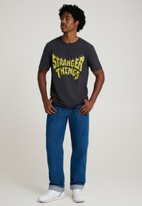 Harmaa T-paita, jossa on keltainen "Stranger Things" -tekstisuunnittelu, yhdistettynä sinisiin farkkuihin ja valkoisiin lenkkareihin, neutraalilla taustalla.