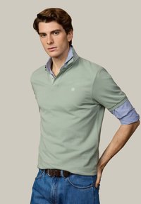 Jeune homme aux cheveux bruns ondulés portant un polo vert superposé à une chemise rayée bleue, associé à un jean bleu et une ceinture tressée marron.