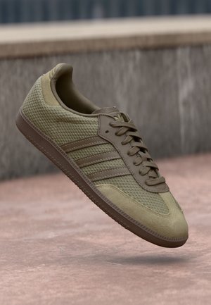 Light brown Adidas sneaker with mesh and suede panels, three stripes on the side, and a textured sole, floating above a pinkish ground.