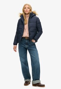Veste en duvet marine avec une capuche en fausse fourrure, associée à un pull en tricot texturé rose et à un jean bleu à jambe large. Des chaussures marron complètent la tenue.