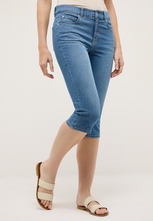 Kvinde iført blå denimshorts i mellemlængde og beige flade sandaler med dobbelt strop, stående mod en ensfarvet baggrund.