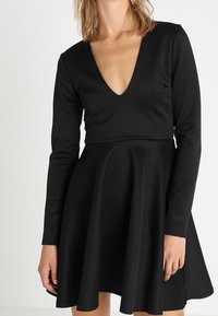 Musta mekko, jossa pitkät hihat ja syvä v-pääntie. Leveä helma, valmistettu sileästä, joustavasta kankaasta. Minimalistinen tyyli ilman kuvioita.