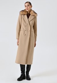 Cappotto beige doppiopetto con grandi bottoni e colletto in pelliccia sintetica, caratterizzato da un design sartoriale e una silhouette lunga e dritta. Stivali neri al ginocchio.