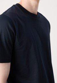 Man som bär en enkel svart t-shirt med kort ärm och rund hals, fotograferad från nacken till mitten av överkroppen mot en enhetlig ljus bakgrund.