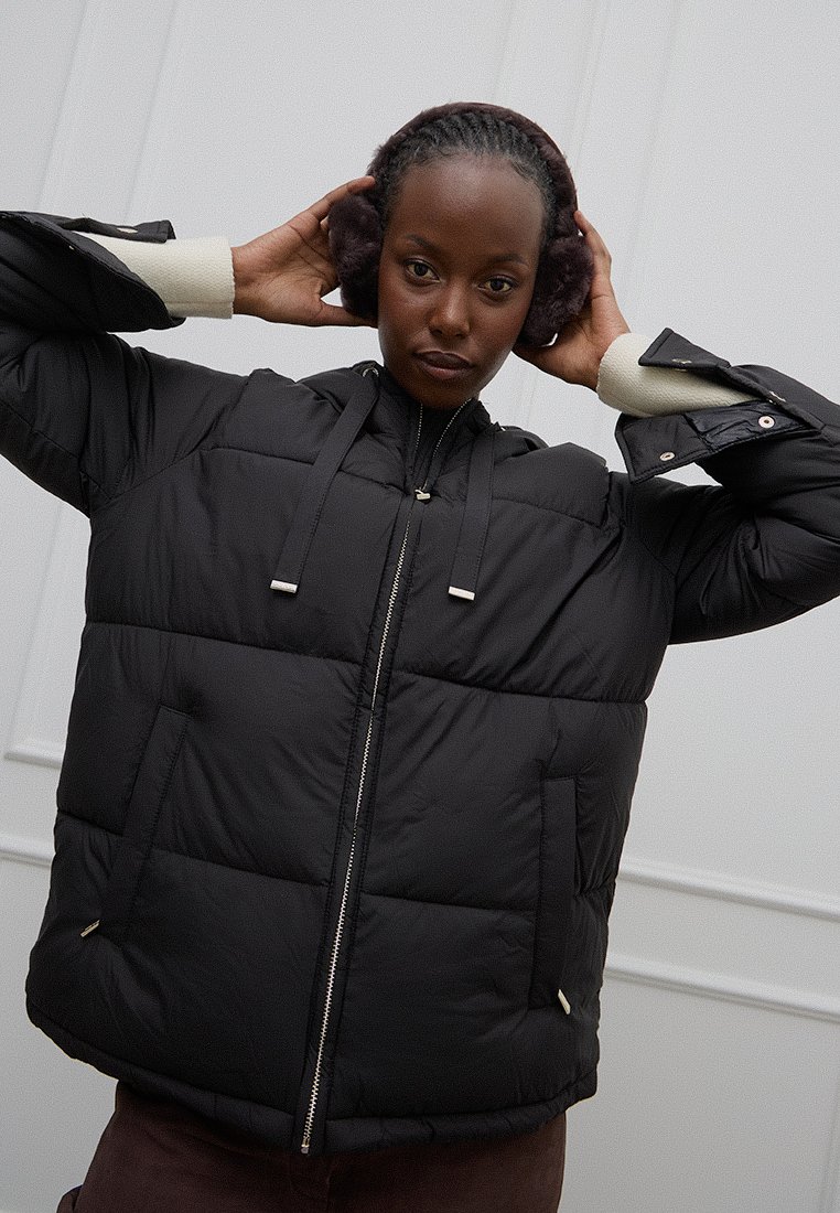 Chaqueta acolchada negra con capucha, con secciones de acolchado horizontal y cierre de cremallera. Acentos crema en los puños y detalles en el herraje.