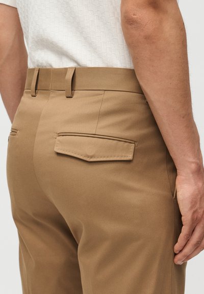 Άνδρας που φοράει καμηλό tailored παντελόνια με πίσω τσέπες με καπάκι και ένα λευκό υφασμάτινο πουκάμισο, εμφανιζόμενος από τη μέση και κάτω.