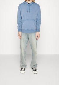 Světle modrá mikina s kapucí a přední kapsou, v kombinaci se světle modrými džíny a černými teniskami s bílou podrážkou. Jednoduchý design, volný střih.