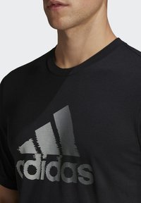 Mustassa puuvillapaidassa on suuri, tekstuurilla varustettu hopeinen Adidas-logo rinnassa. Pyöreä pääntie ja lyhyet hihat.