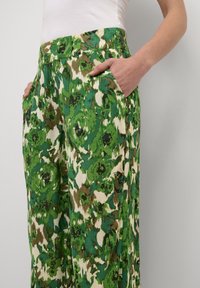 Pantaloni a fantasia floreale verde con una combinazione di tonalità verde chiaro e scuro, crema e marrone. Presentano tasche e una vita piatta.