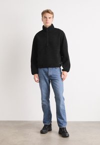 Zwarte fleece pullover met een hoge kraag en een gedeeltelijke rits, gecombineerd met lichtblauwe spijkerbroek en zwarte veterschoenen.