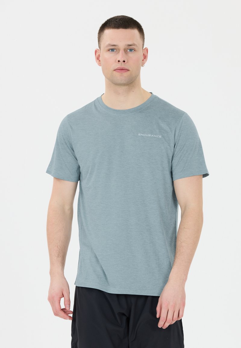 Mies, jolla on päällä vaalea sininen lyhythihainen T-paita, jossa on teksti "ENDURANCE", ja mustat shortsit, seisoo valkoista taustaa vasten.