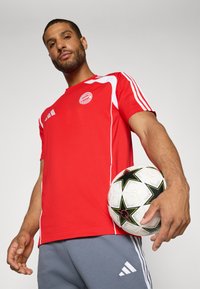 Red athletic T-shirt with white accents and logo, paired with gray shorts. The model holds a black and white soccer ball with star patterns.