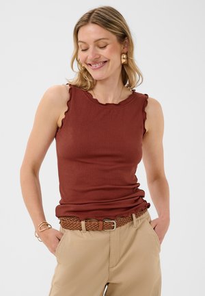 Femme aux cheveux blonds portant un haut marron sans manches côtelé et un pantalon beige avec une ceinture tressée, souriant les yeux fermés et mains dans les poches.