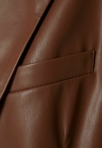 Brązowa sztuczna skóra o delikatnej, ziarnistej fakturze, z eleganckim, skośnym detalem kieszeni. Powierzchnia ma lekki połysk i głębię.