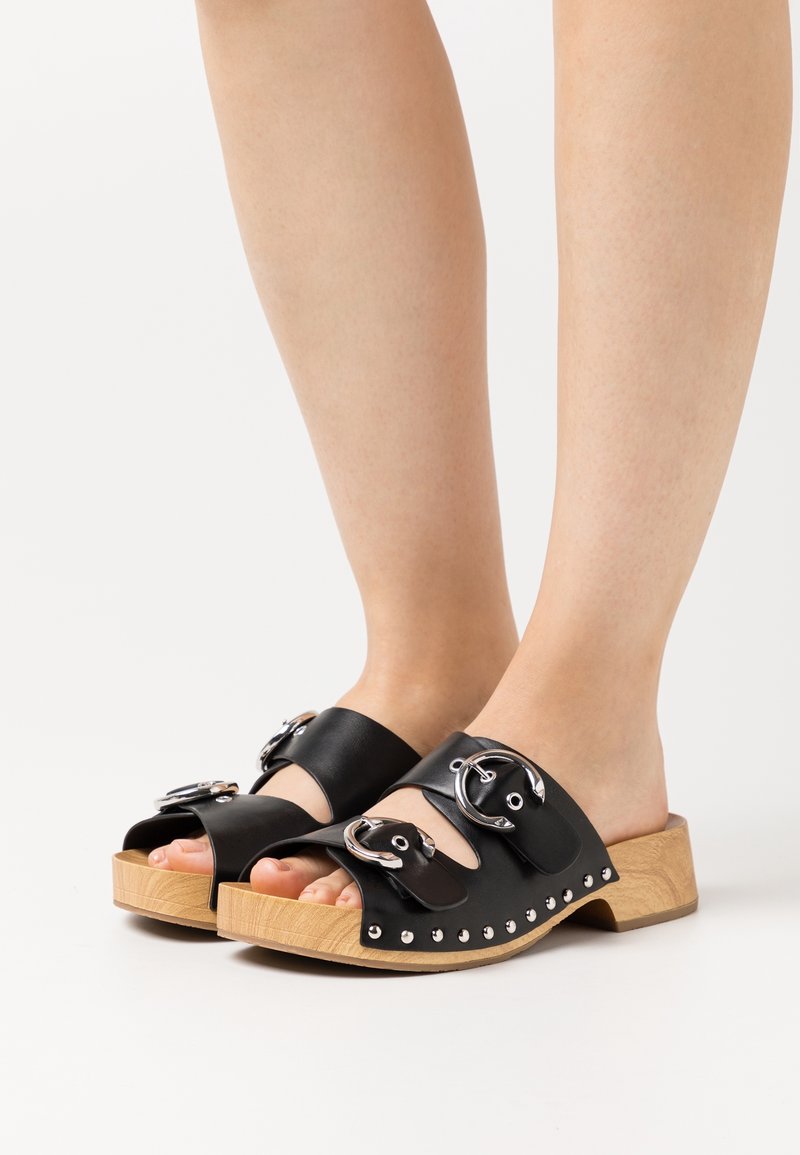 Pieds portant des sandales en cuir noir à enfiler avec des boucles et des clous argentés, et des talons blocs bas en bois sur fond blanc.