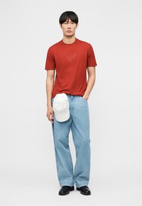 Muž nosiaci červenú košeľu s krátkymi rukávmi, svetlomodré široké rifle, čierne topánky, drží bielu šiltovku a má ruku v vrecku.