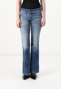 Jeans a zampa d'elefante in denim blu con una lavatura sbiadita, tasche frontali e chiusura a doppio bottone. Abbinati a scarpe nere con tacco.