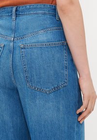 Person som har på seg høytlivde blå denimjeans med synlig baklomme og bar høyre arm mot en hvit bakgrunn.