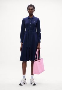 Vestito lungo blu navy con maniche lunghe, chiusura con bottoni davanti e cintura in vita, abbinato a una borsa a tracolla rosa, sneakers bianche e calzini a righe.