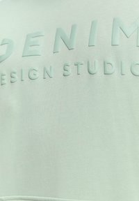 Felpa color verde menta chiaro con lettering in rilievo "DENIM DESIGN STUDIO." Tessuto morbido e testurizzato con un'estetica pulita e moderna.