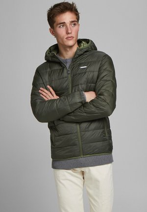 Grüne Pufferjacke mit Kapuze, Reißverschluss und gestepptem Design. Darunter trägt die Person einen grauen Strickpullover und cremefarbene Hosen.
