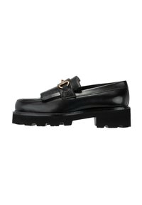 Mocasín de cuero negro con suela gruesa y estriada, detalle de flecos y hebilla en tono dorado sobre fondo blanco.